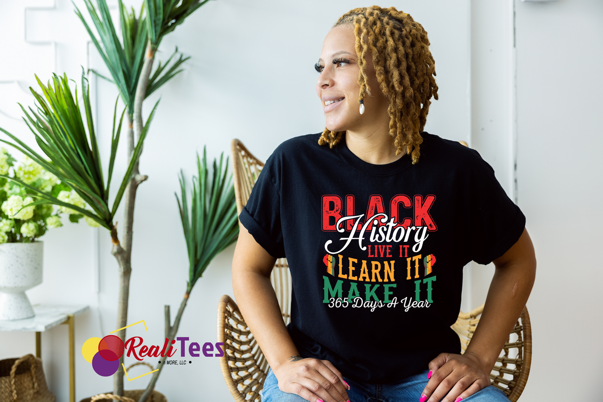 Person wearing a black t-shirt with 'Black History Live It Learn It Make It 365 Days A Year' text, sitting in a room with plants.